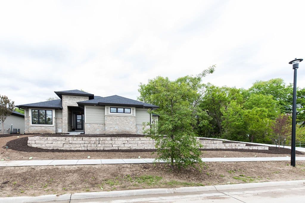 Modern single-story home with a mix of stone veneer and horizontal siding exterior. The property features a dark gabled roof, a retaining wall with landscaping, and a well-manicured lawn.