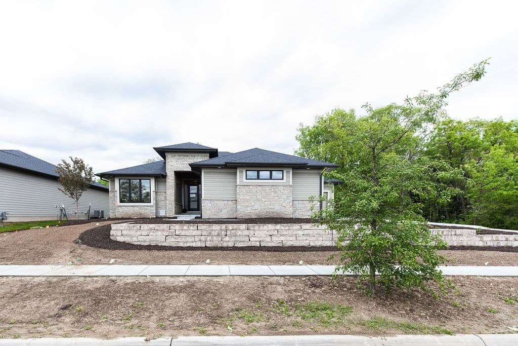 Modern single-story home showcasing a stone veneer facade and light gray horizontal siding. The professionally landscaped yard features a tiered retaining wall and young trees.