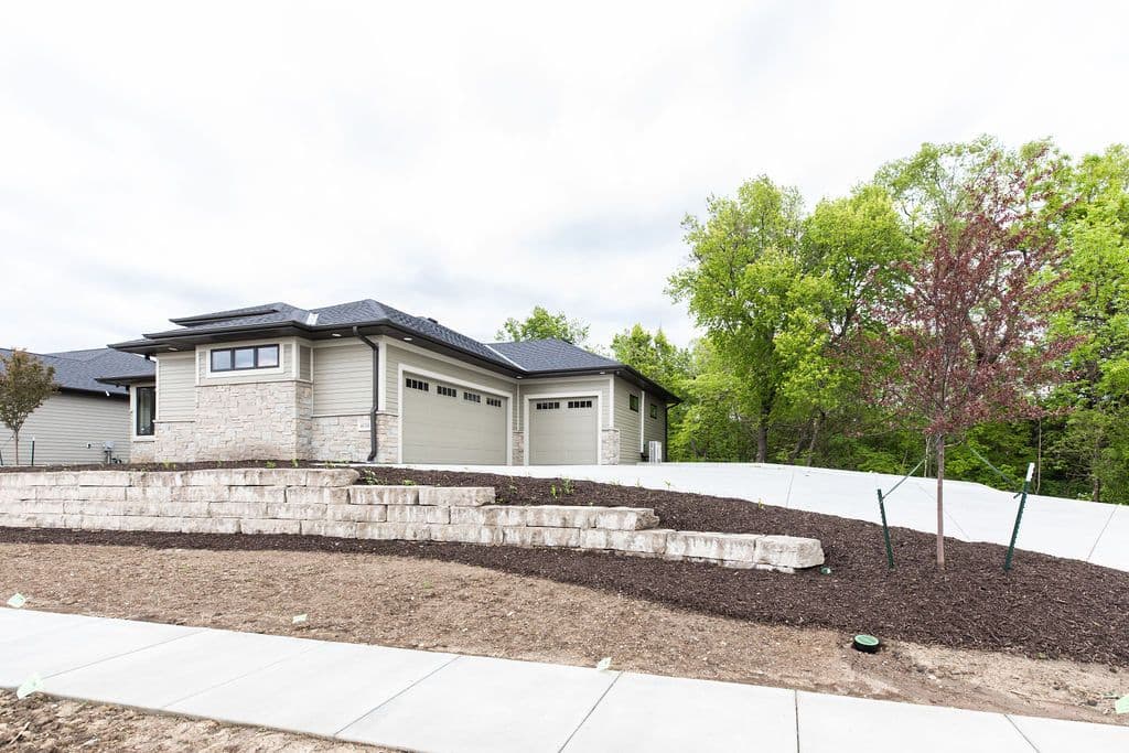Exterior view of a modern home featuring a two-car garage, light beige horizontal siding, and a stone veneer accent wall. A tiered retaining wall and landscaped garden bed are visible in the foreground.