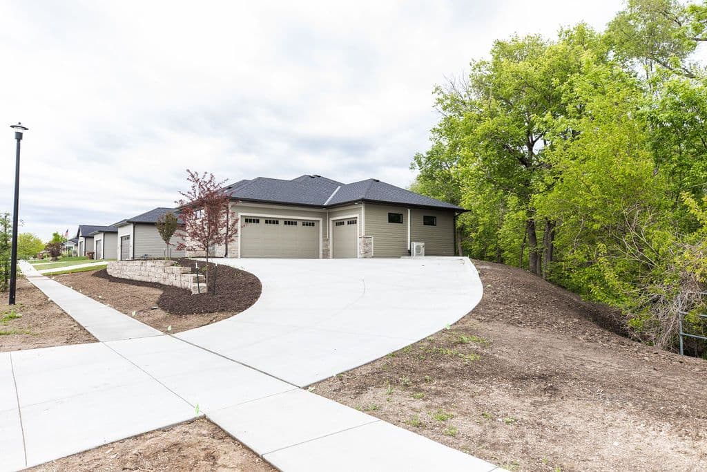 Exterior view of a modern home with a concrete driveway leading to a two-car garage. The home features gray siding and a dark gray roof, with green trees in the background and a stone retaining wall.