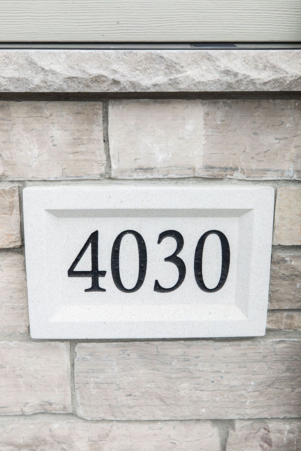 Close-up of a rectangular stone address plaque displaying the numbers "4030" in bold black font. The plaque is mounted on a light brown stone wall with gray horizontal siding visible above.