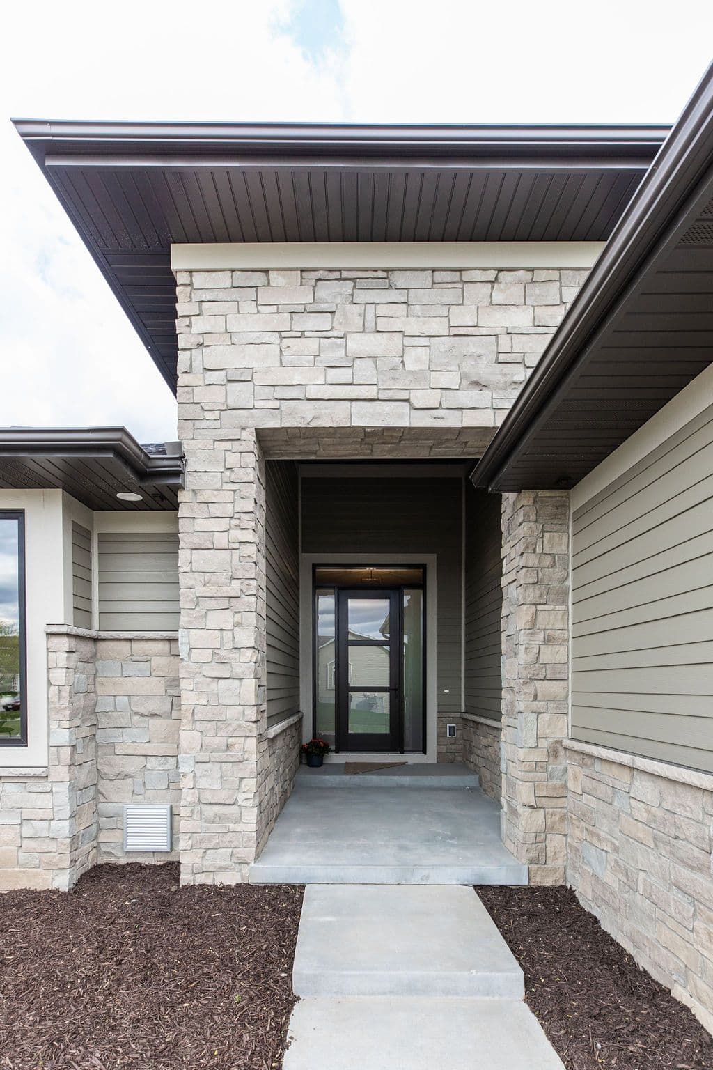 Exterior of a modern home featuring a stone veneer entrance and horizontal siding, a brown roof, and a concrete walkway leading to the front door. The door is glass and framed in black, set within a recessed entryway.