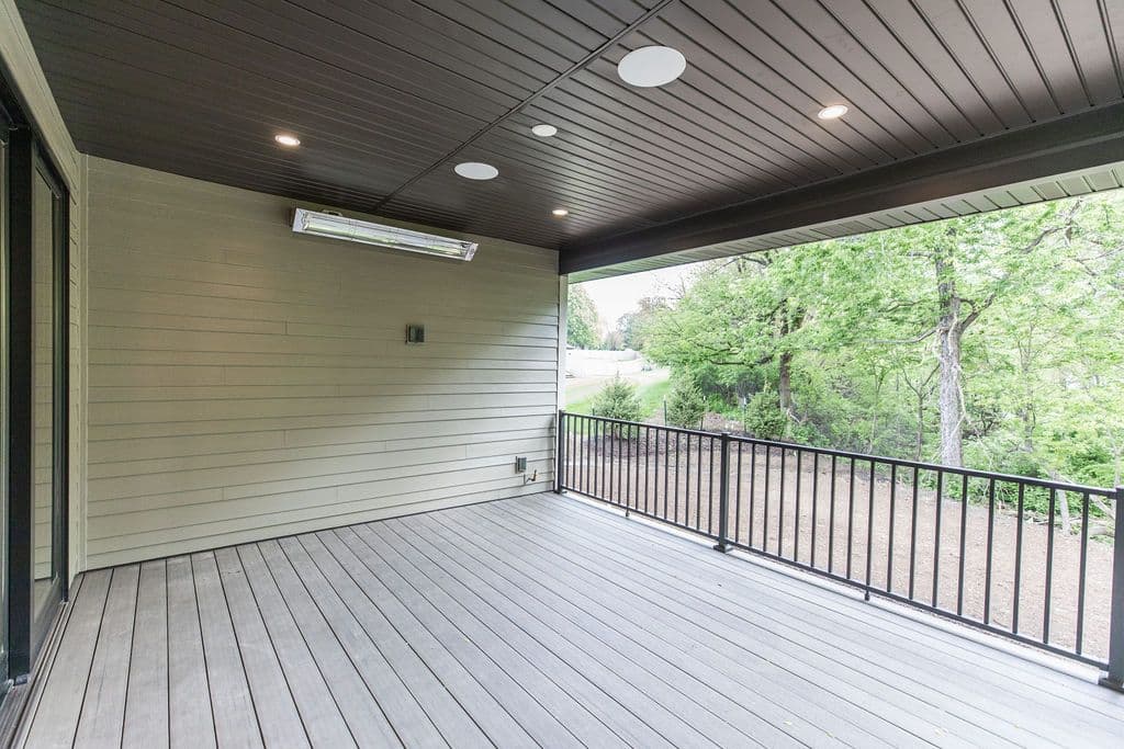 Covered outdoor deck featuring composite deck boards and a black metal railing overlooking a wooded yard. The space is lit by recessed lighting and heated by a wall-mounted heater.