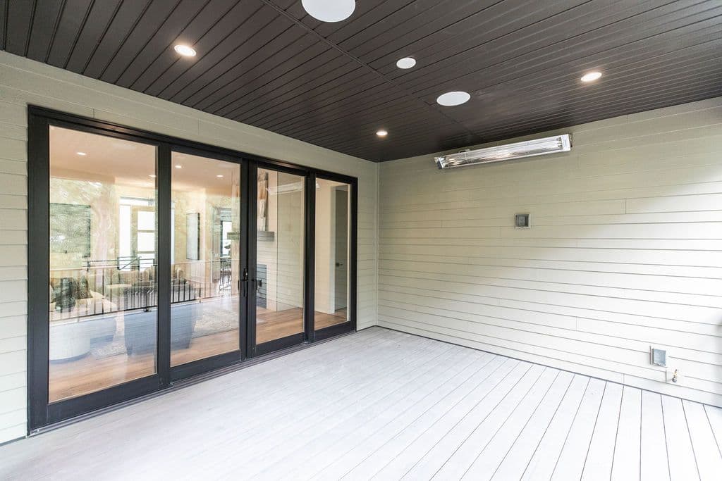 Covered patio with black framed sliding glass doors leading to the interior. The outdoor area features a light gray deck, horizontal siding, a dark gray ceiling with recessed lighting, and a mounted heat lamp.