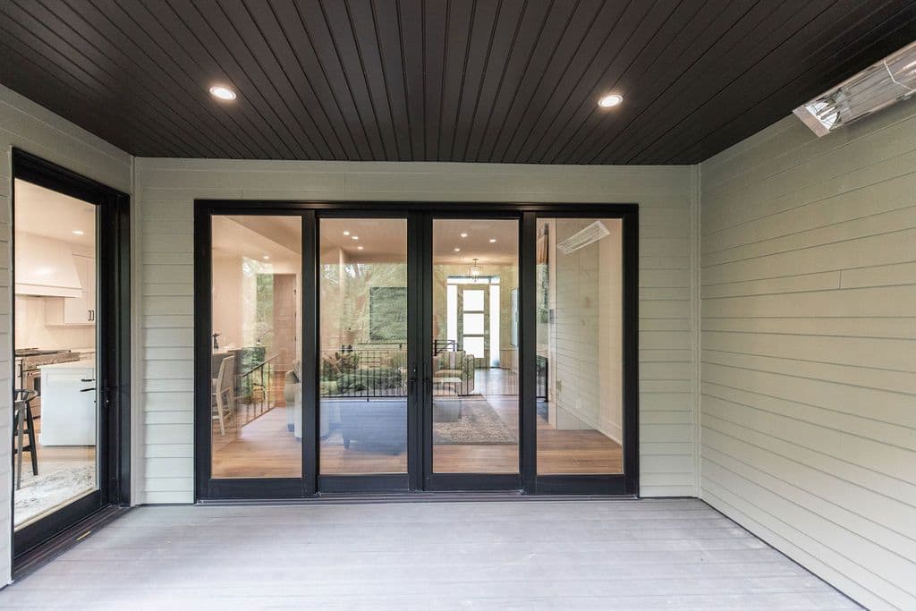 Covered outdoor patio featuring black-framed sliding glass doors leading to the interior. The patio has a gray wood floor and is sheltered by a dark-stained wood ceiling.