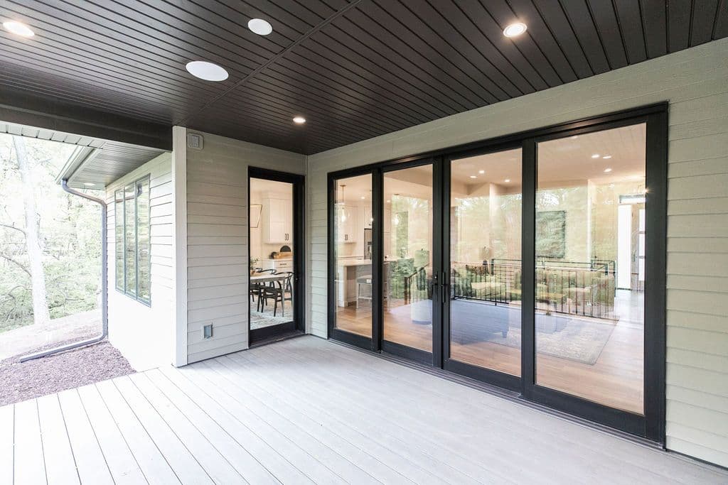 Covered patio featuring a light gray wood deck and horizontal siding. Black framed sliding glass doors provide access to the interior while recessed lighting illuminates the space.