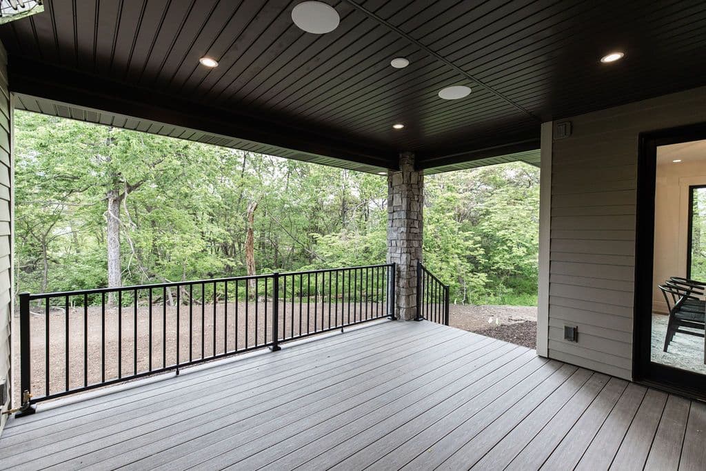 Covered outdoor deck with a black railing overlooks a green, wooded area. The deck has a grey wood floor and a stone support pillar, with a sliding glass door leading into the house.