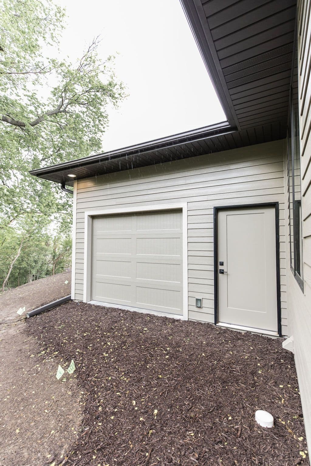 Exterior view of a one-car garage with a side entry door and horizontal siding. The area around the garage is covered in dark brown mulch.