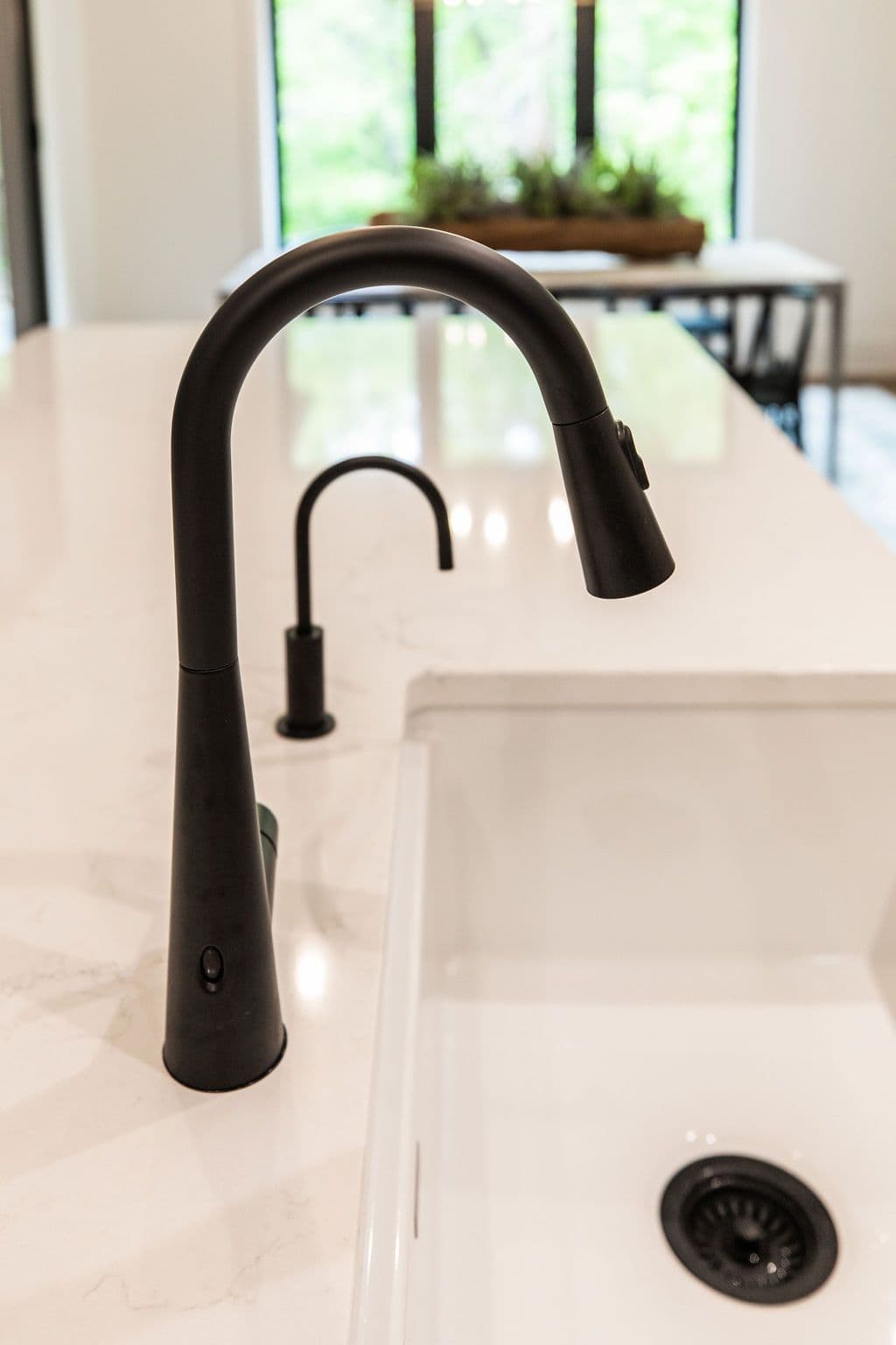 Close-up shot of a modern kitchen sink with a sleek matte black faucet and white countertop with subtle marbling. The sink is also white, creating a clean and minimalist look.