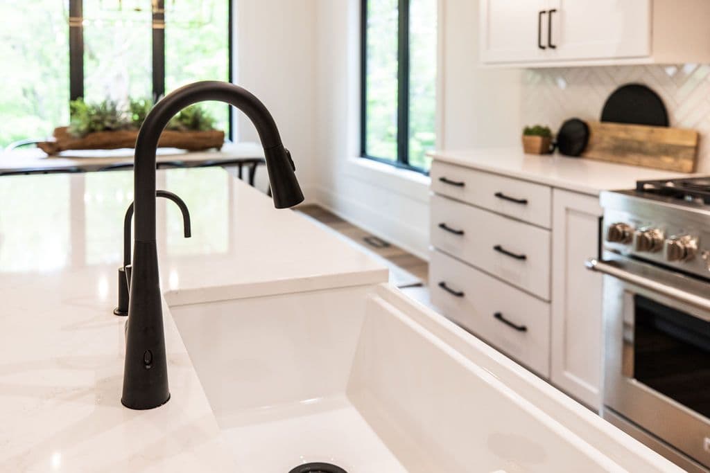 Modern kitchen features a matte black faucet and white farmhouse sink set in a marble-patterned countertop island. White cabinets and stainless steel oven and range are visible in the background, along with a herringbone backsplash.