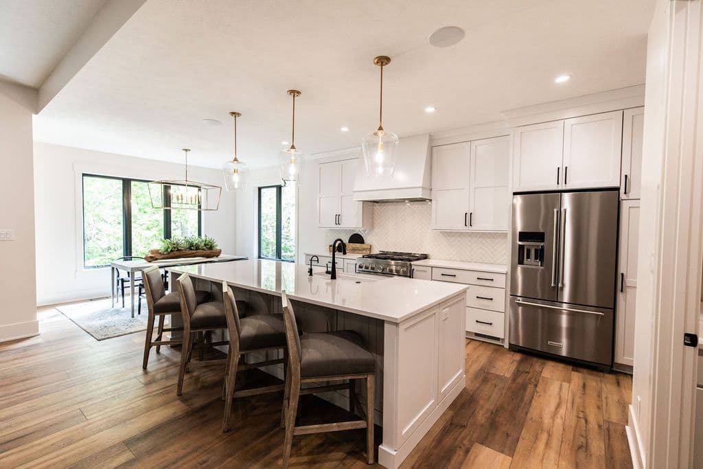 Bright, modern kitchen features white cabinets, stainless steel appliances, and a large central island with seating. The adjacent dining area has a dark-framed window and contemporary light fixture.