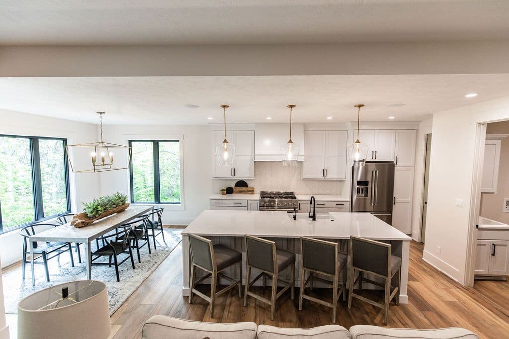 Bright, open kitchen and dining area with white cabinets, a large island with seating, and black framed windows providing ample natural light. The dining table features a rustic centerpiece on a patterned area rug.