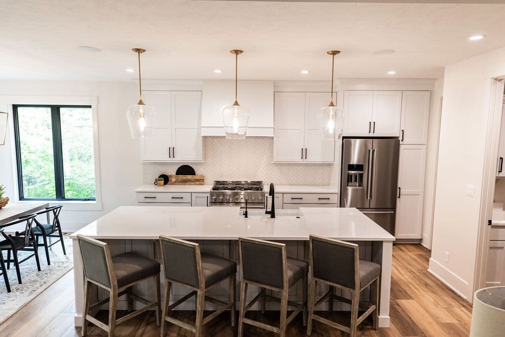 Modern kitchen with white shaker cabinets, a large white island with barstool seating, and stainless steel appliances. Three pendant lights hang over the island and the kitchen features hardwood flooring.