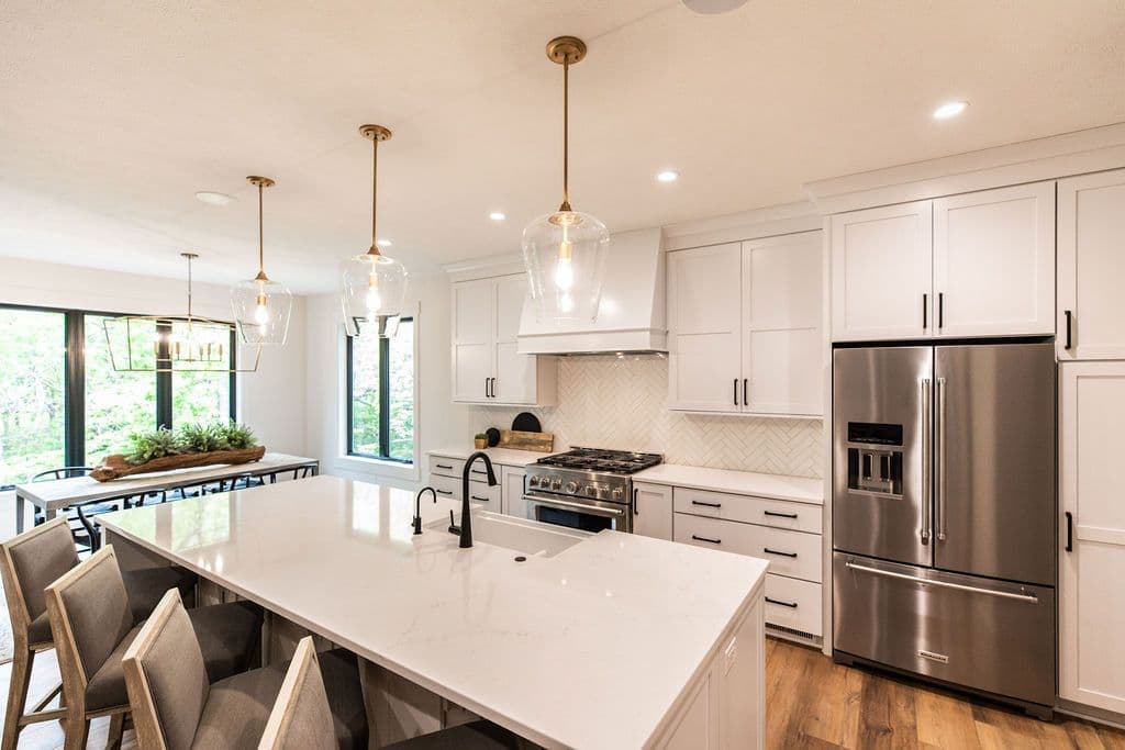 Bright and modern kitchen featuring a large white marble island with seating, white cabinetry with black hardware, and stainless steel appliances. The space is illuminated by pendant lighting with glass shades and has wood flooring.