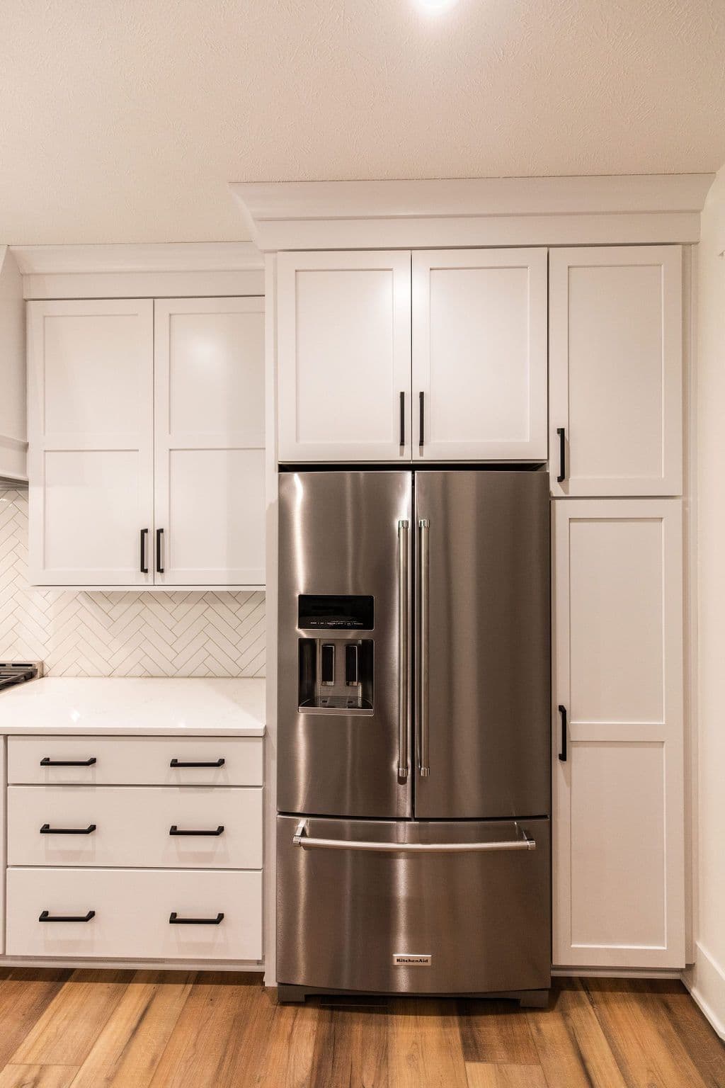 Modern kitchen features stainless steel refrigerator with white cabinetry and black hardware. The floor is hardwood and there is a white countertop with a herringbone backsplash.