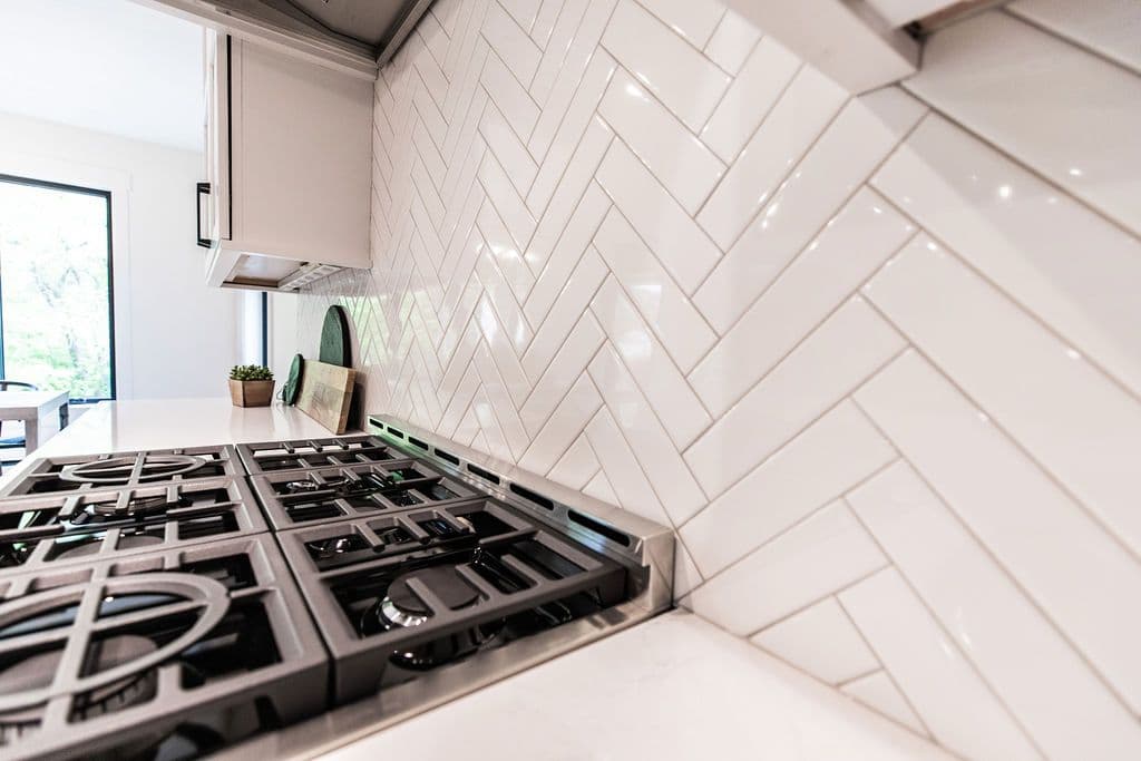 A modern kitchen showcases a gas stovetop with stainless steel trim and a white countertop. The backsplash is composed of white subway tiles laid in a herringbone pattern.