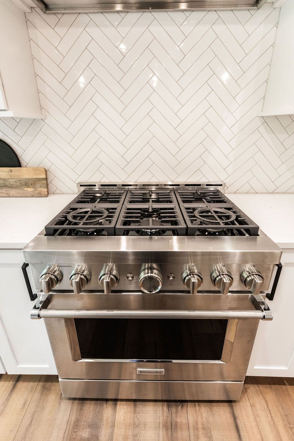 Stainless steel KitchenAid range with six burners and white herringbone tile backsplash. The oven is also stainless steel and the floor is light brown hardwood.