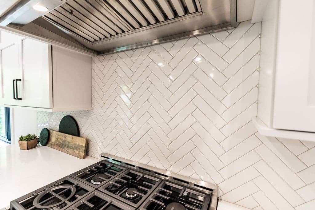 Contemporary kitchen with white herringbone tile backsplash, stainless steel range hood, and white cabinetry. The gas stovetop has black grates, contrasting with the light countertop.