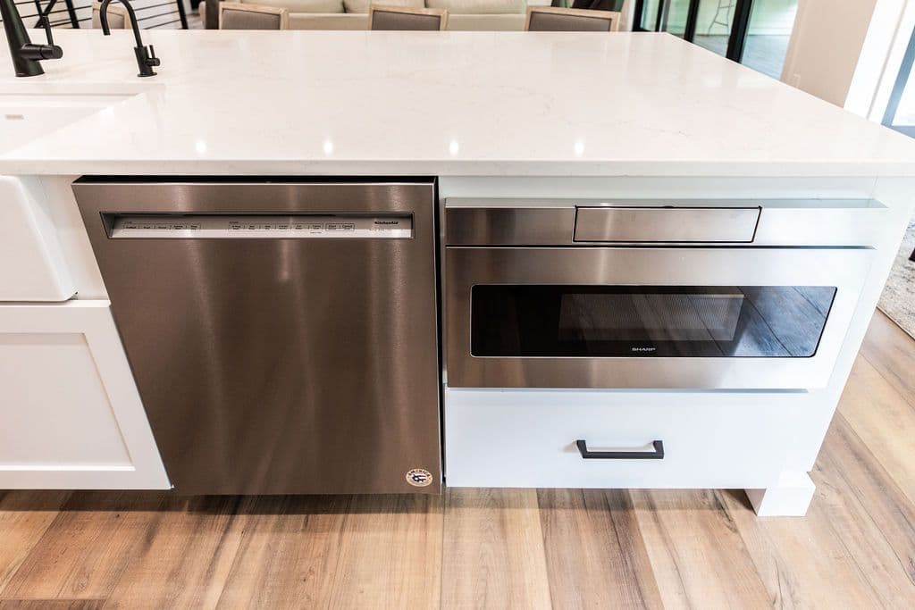 Close-up of a kitchen island featuring a stainless steel dishwasher and a drawer microwave beneath a white countertop. The cabinets are white with simple black drawer pulls.