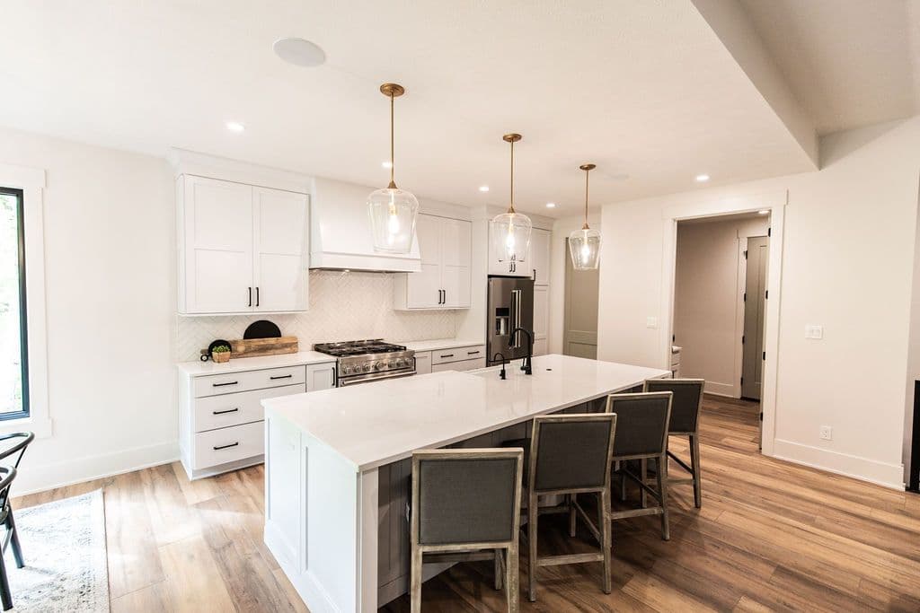 Bright kitchen featuring a large island with bar stool seating, white cabinets with black hardware, and stainless steel appliances. Three pendant lights with clear glass shades hang over the island, complementing the warm wood flooring.