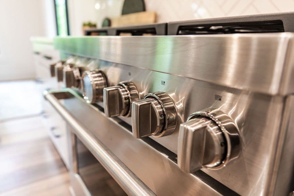 Stainless steel range with multiple knobs and burners in a kitchen setting. The range features a modern design with a handle and oven door visible, surrounded by light-colored surfaces.