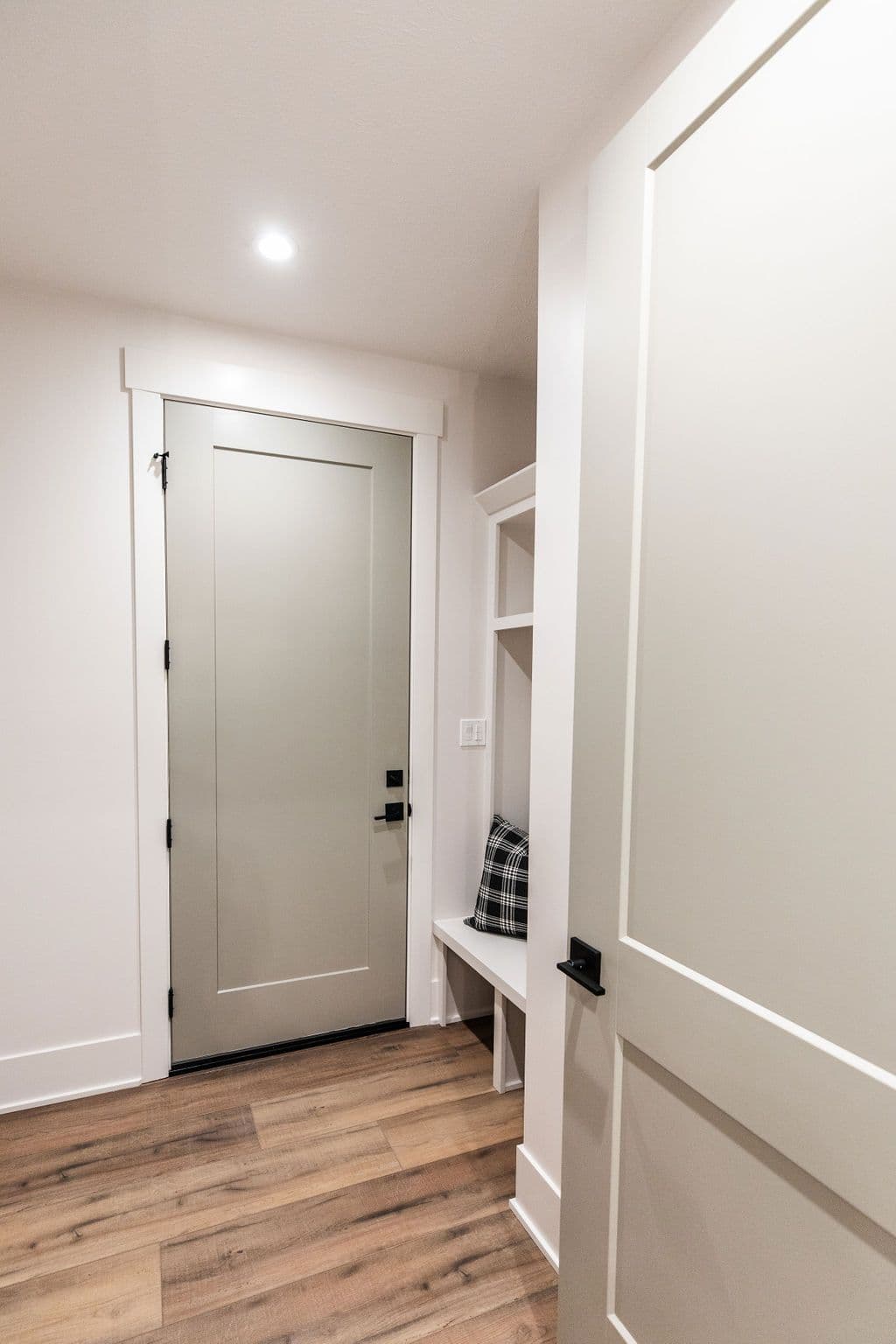 Entryway with a gray door and white trim, a built-in bench and shelf with a plaid pillow, and wood-look flooring. A white door on the right side is partially visible.