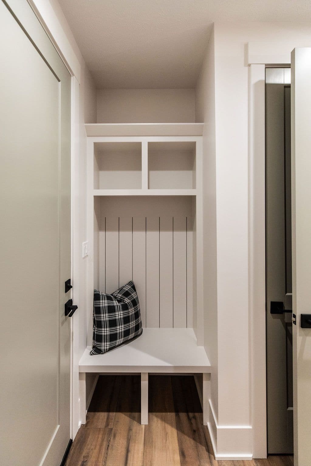Entryway mudroom nook features built-in white shelving and bench seating with a black and white plaid pillow. Hardwood floors run throughout the hallway near the mudroom area and a contrasting dark door.