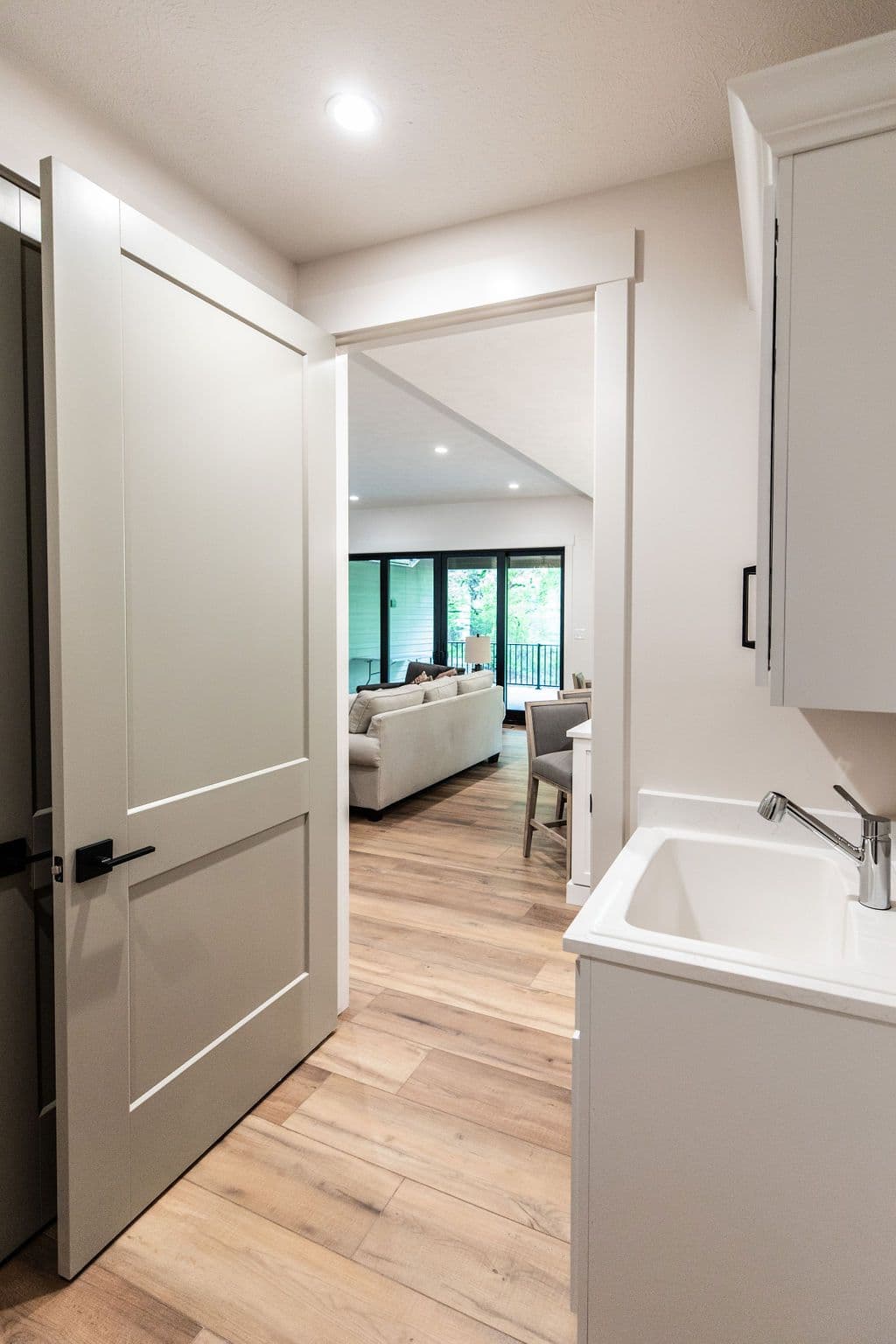 Interior view showcases a hallway with an open doorway leading into a living room. A white sink cabinet is visible in the foreground, with light wood flooring extending through the hallway and living room.