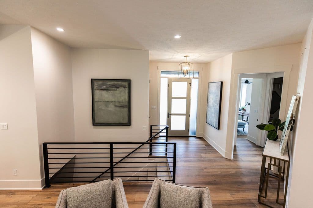 Bright home interior featuring hardwood floors, modern black railing, and neutral decor. Art hangs on the walls, with a view into a front office and a door leading outside.