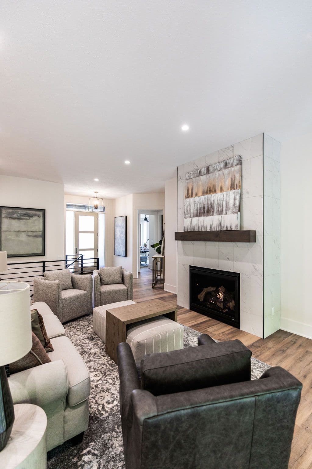 Bright living room with a fireplace featuring white marble tile and a dark wood mantel. The room contains neutral-toned furniture, including a sofa, chairs, and ottoman, arranged on a patterned rug.
