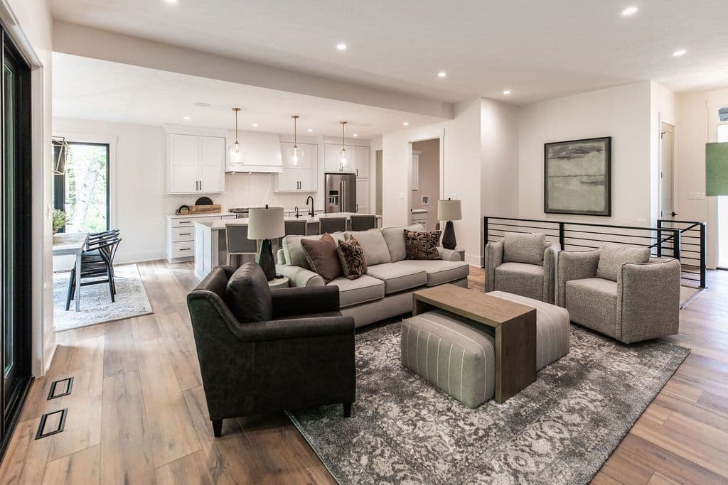 Open concept living room and kitchen with hardwood floors and recessed lighting. The living room features a gray sofa, two armchairs, and a gray patterned rug, while the kitchen has white cabinets and a stainless steel refrigerator.