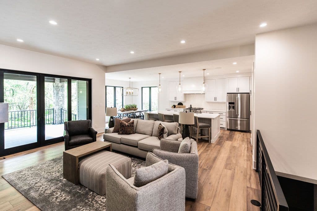 Open concept living space features living room, dining area, and kitchen. The living room has gray furniture, a patterned rug, and black framed sliding glass doors. Kitchen features white cabinetry, stainless steel appliances, and an island with bar seating.