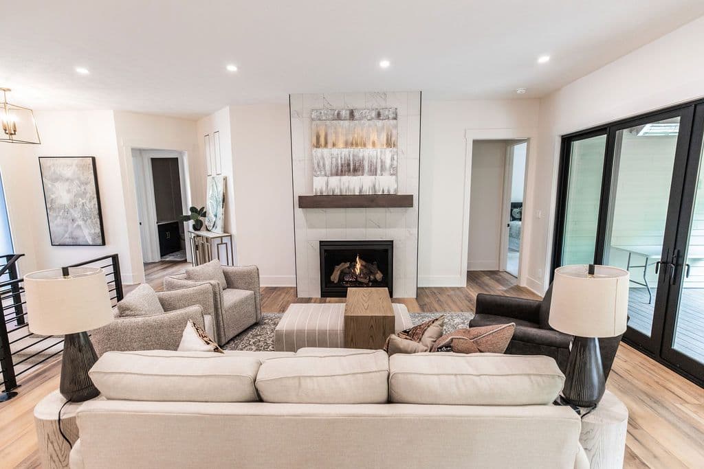 Bright living room with a fireplace feature wall clad in marble tiles and topped with a wooden mantle. Neutral-toned furniture including a sofa, armchairs, and upholstered ottomans center the space with wood floors and views to the exterior.