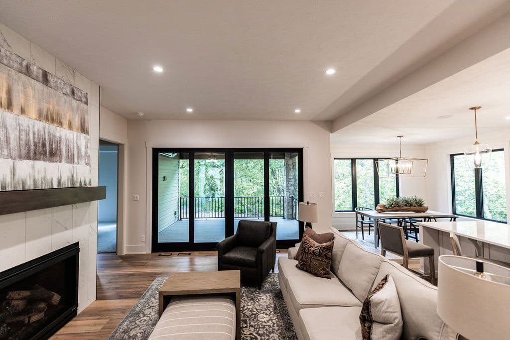 Open concept living space with fireplace, modern furniture, and sliding glass doors leading to a deck. Dining area with large windows is visible adjacent to the living room.