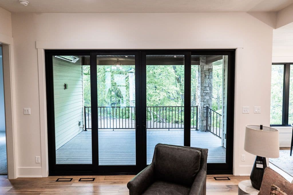 Interior shot of a room with a four-panel black frame sliding glass doors leading to an outdoor deck with a black railing. A gray upholstered armchair sits in the foreground.