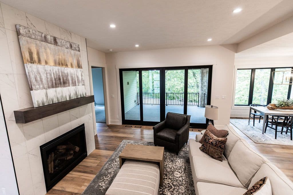 Modern living room with a fireplace featuring a neutral painting and a dark wood mantle. The room has hardwood floors, a gray patterned rug, and large sliding glass doors leading to an outdoor deck, with a glimpse of a dining area to the right.