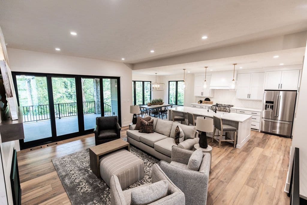 Open concept living and dining area featuring a gray sofa and armchairs on a patterned rug, and a dining table and kitchen with a large island and stainless steel appliances. Expansive windows with black frames provide natural light and views of the outdoors.