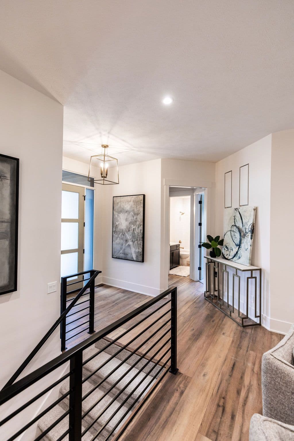 Hallway featuring wood floors leads to a bathroom and artwork-adorned console table. Black metal railing overlooks a staircase with carpet.