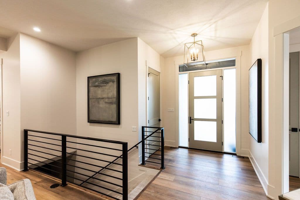 Modern entryway with wood floors, a geometric chandelier, and an abstract painting on the wall. A black metal railing leads to a lower level.