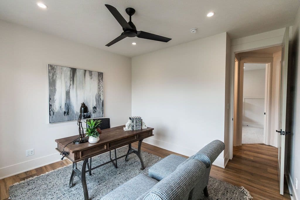 This office space features a gray and white abstract art piece above a rustic wooden desk with a black lamp and a small potted plant. A gray patterned rug sits under the desk and a gray chair is in the foreground.