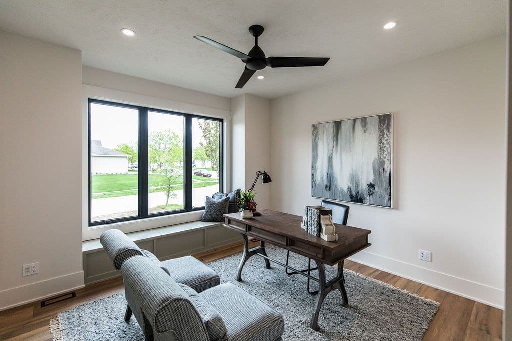 Modern home office with large windows, a built-in window seat with pillows, and a dark wood desk on a gray rug. There is a black ceiling fan and a black and white abstract art piece on the wall.