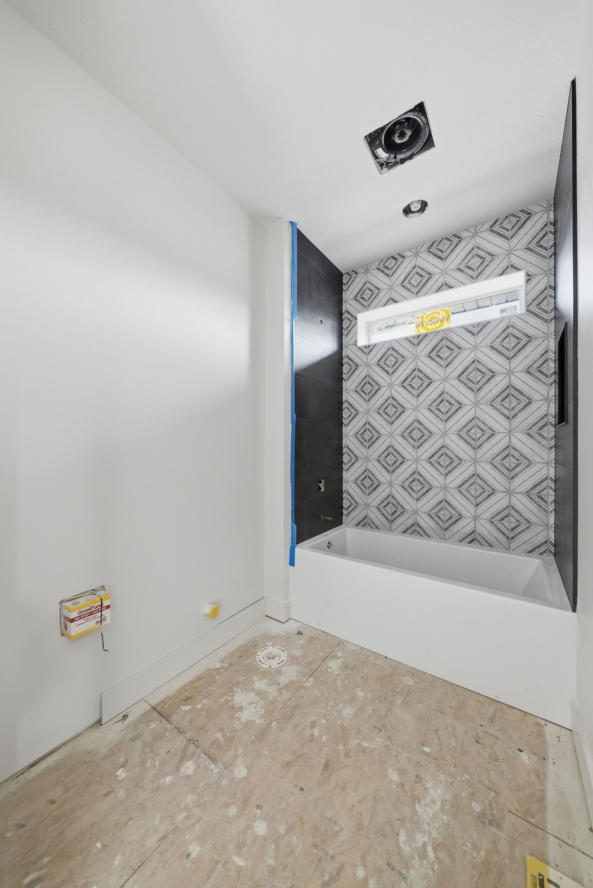 An unfinished bathroom is shown with a modern tiled wall around the bathtub and black accent tiling. The floor is unfinished and there are visible electrical boxes and plumbing stubs.