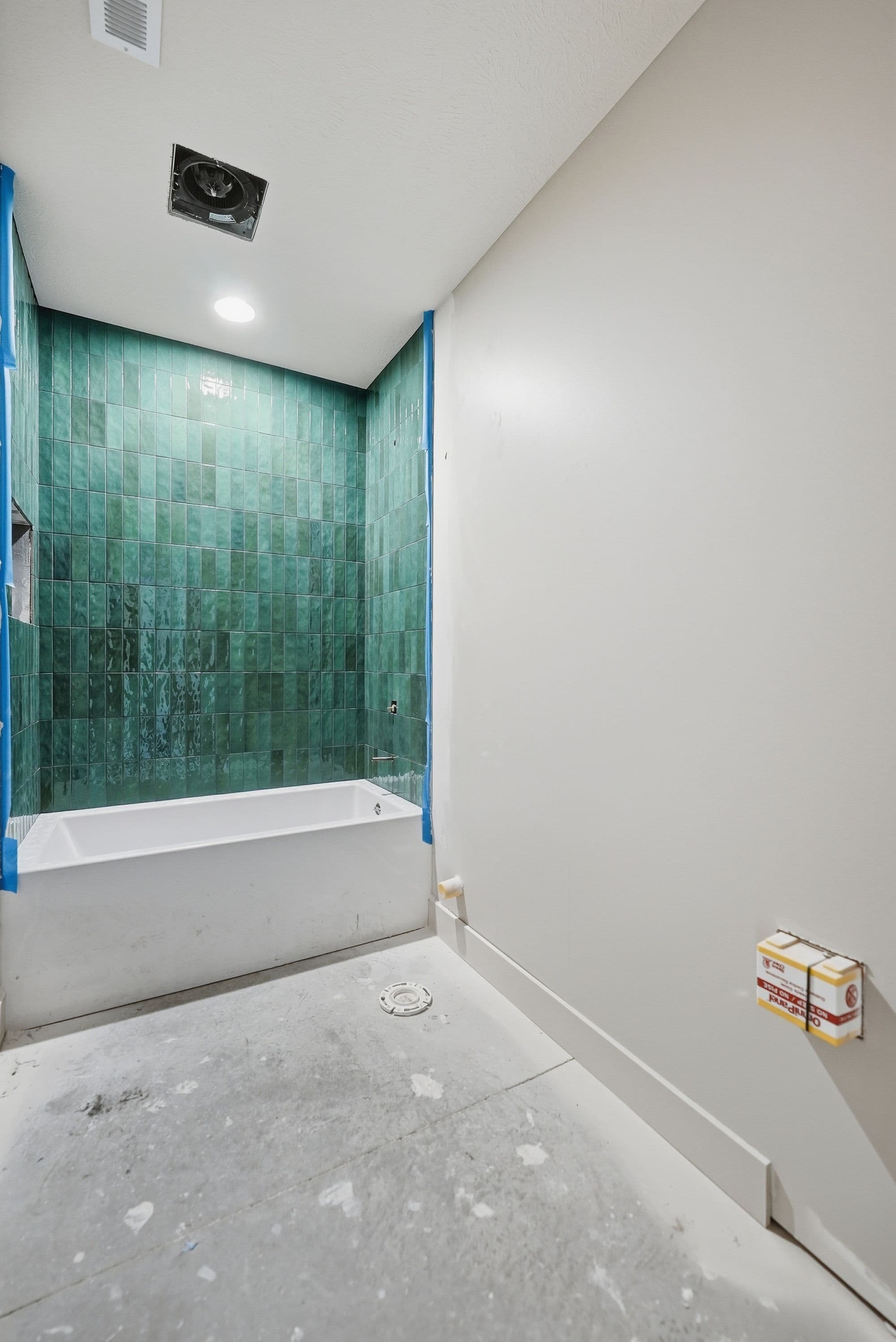 Bathroom under construction showcasing a white bathtub with green tile surround and a modern ceiling fan. The room has a light-colored concrete floor and white walls with white trim.