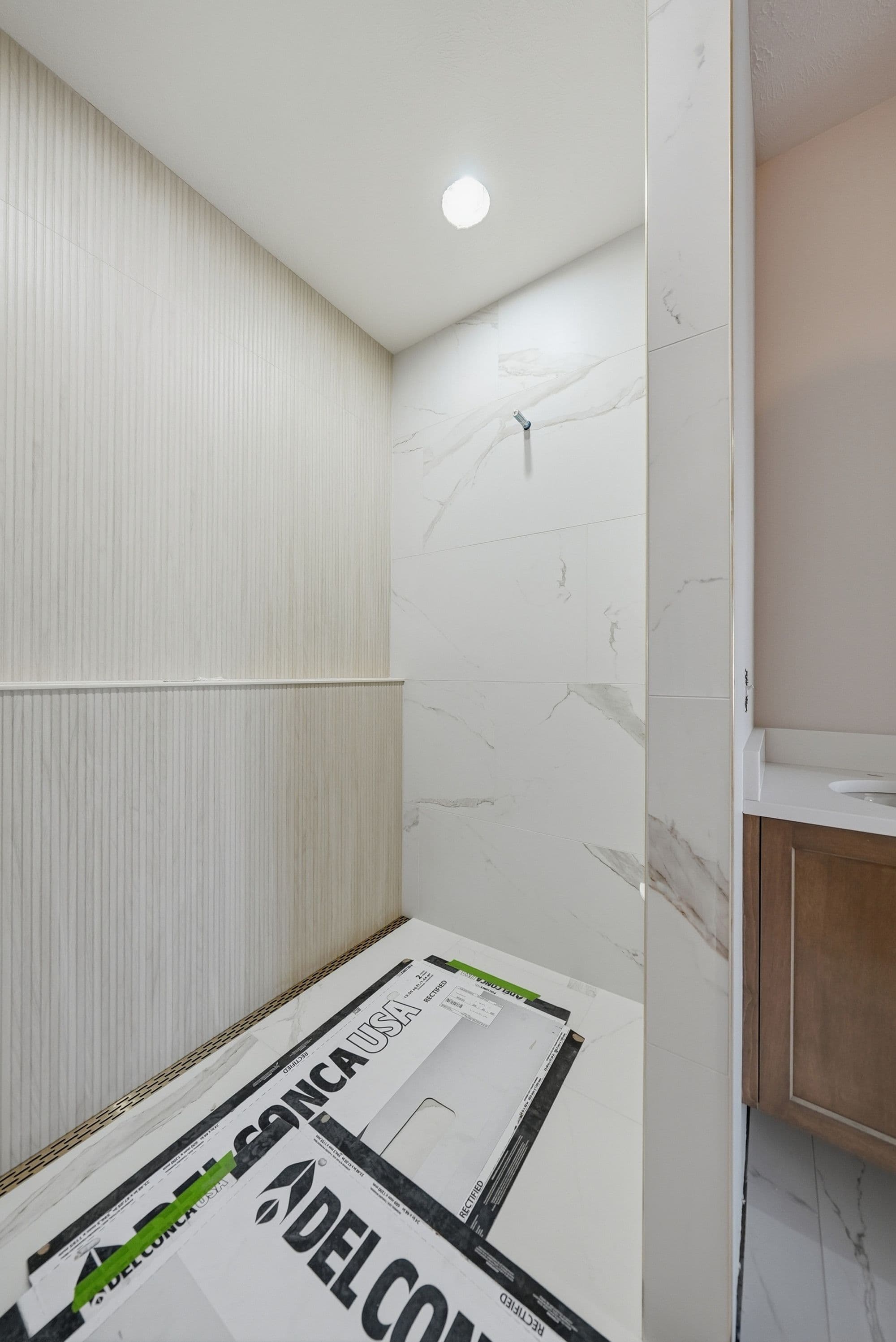 Modern bathroom features horizontally striped wall tiles, marble-look shower tiles, and a recessed ceiling light. Boxes of tile sit on the shower floor, indicating ongoing installation.