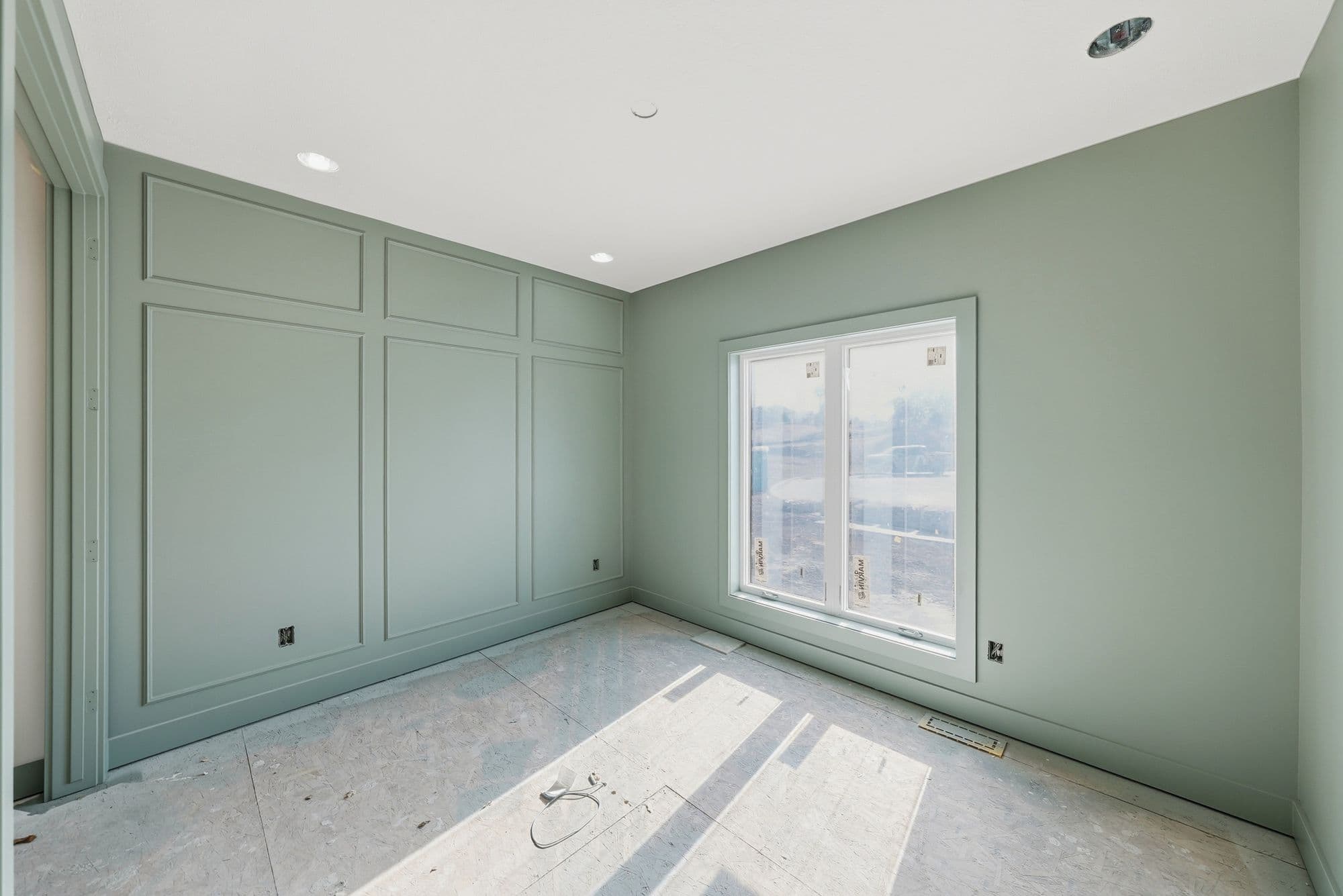 An unfinished room is shown with green walls, white trim, and a large window. The room features decorative paneling on one wall, and the floor is covered with plywood.
