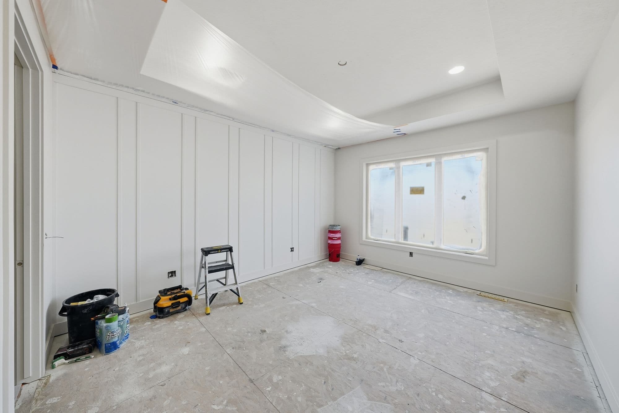 A bright, unfinished bedroom is staged for painting, featuring white paneled walls, a window covered in protective plastic, and a covered ceiling. Painting supplies and a small ladder are scattered around the room.