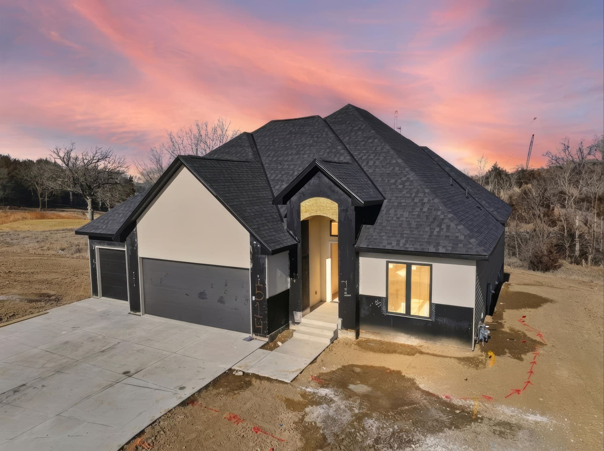 Exterior view of a new construction home featuring a dark gray shingled roof, black painted trim, and light beige siding panels. A concrete driveway leads to the garage, and the yard is bare earth.