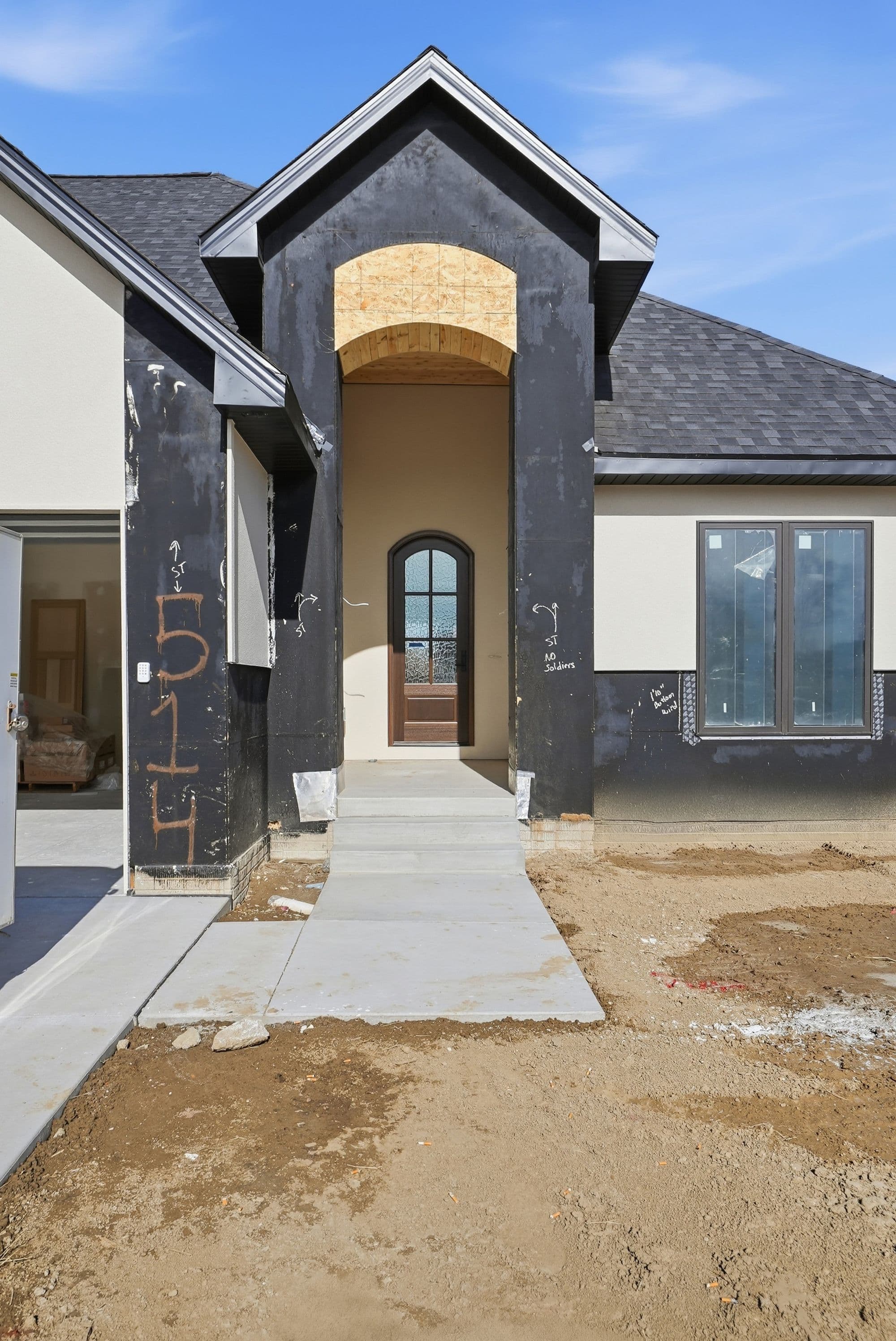 Exterior view of a two-story home under construction. Features include a dark shingle roof, black-wrapped front entrance, stucco siding, and a concrete walkway.