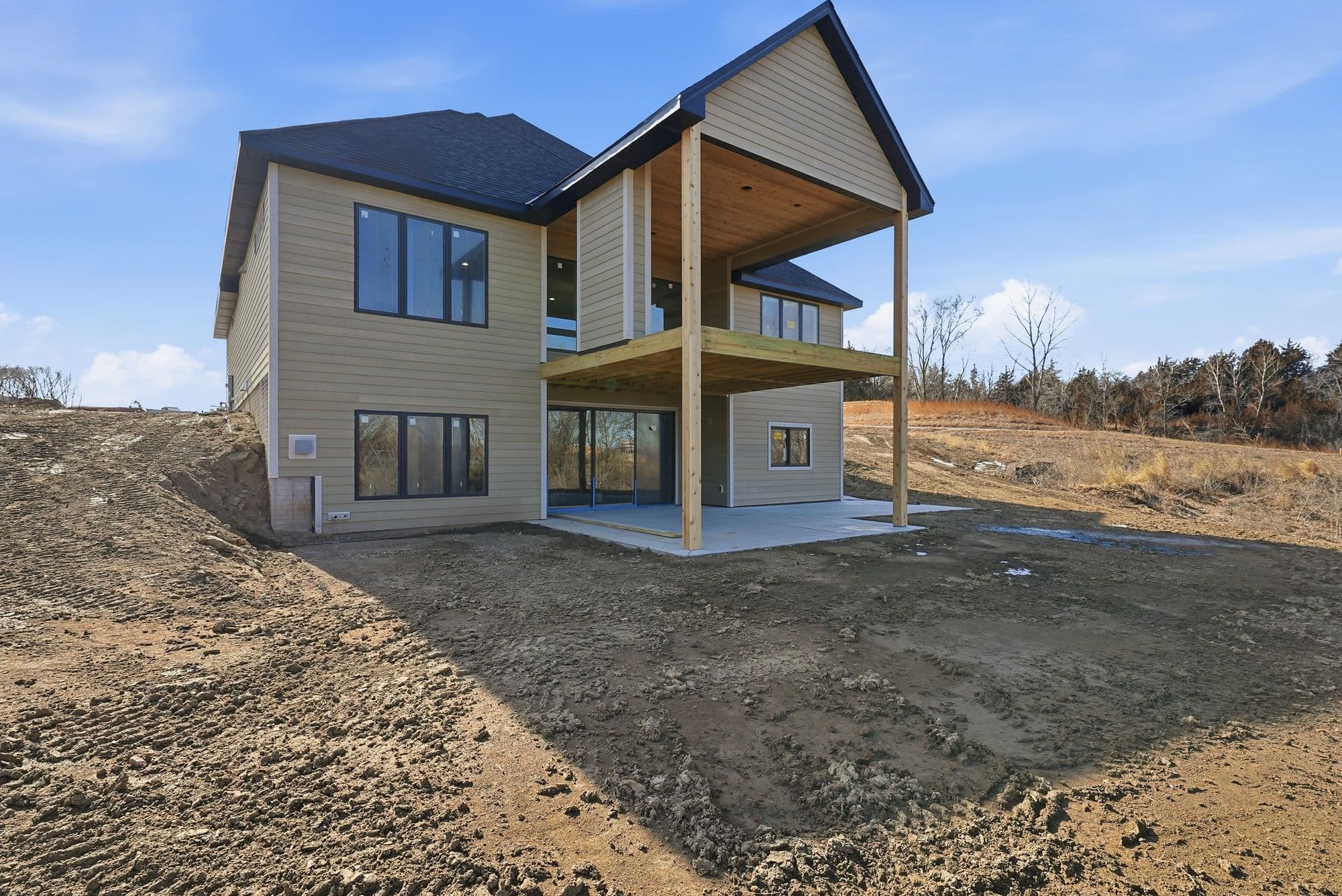 Two-story house featuring a two-story deck with wooden posts and beam construction. The exterior is finished with light brown siding, black framed windows, and a dark roof.