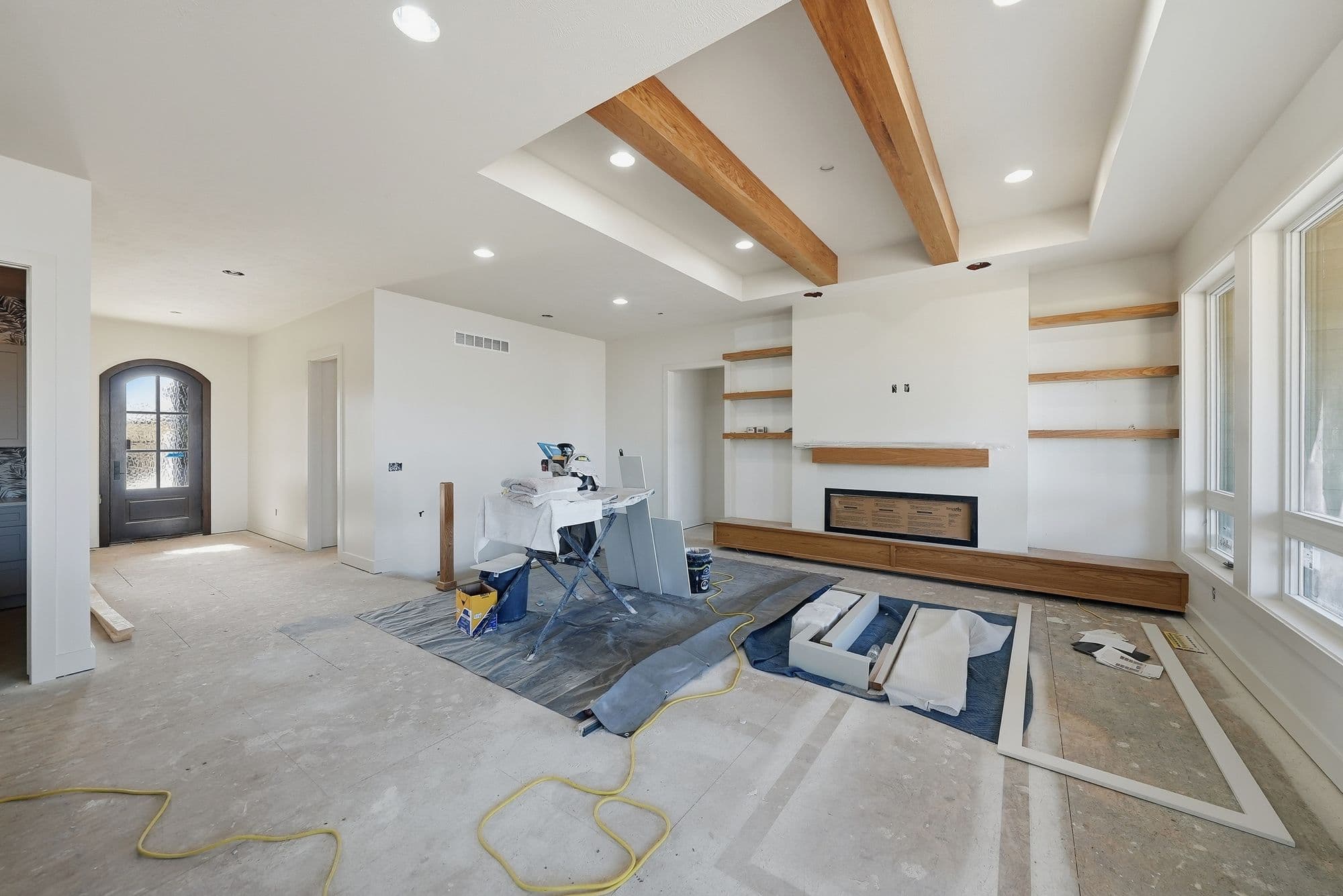 Living room under construction with exposed ceiling beams, built-in shelving, and a modern fireplace. Construction materials and tools are scattered throughout the room.